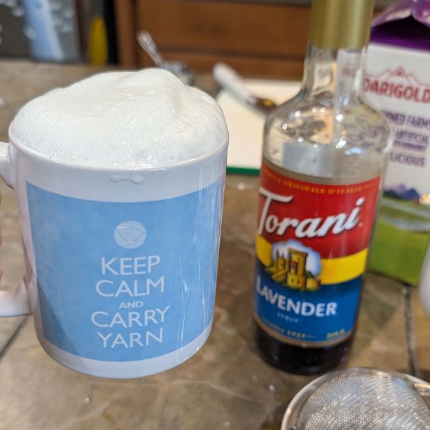 A knitting mug full of London Fog tea with frothed milk on top, next to a bottle of Torani Lavender syrup