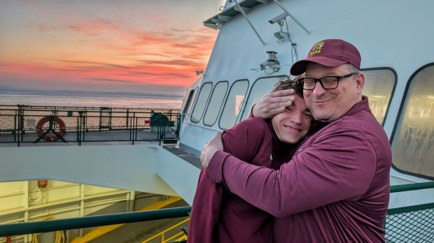 Father and son hug on a ferry boat with a red and orange sunrise in the background